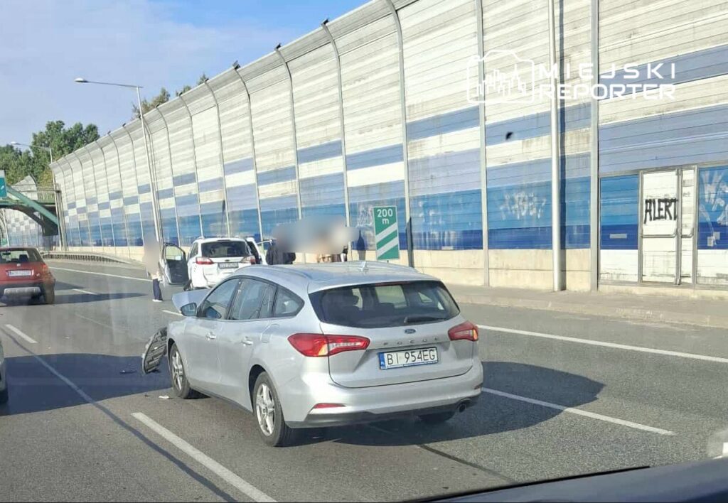 Na jezdni stoi uszkodzony srebrny samochód osobowy, obok niego zatrzymane inne pojazdy. W tle widoczna niebieska ściana akustyczna oraz znak drogowy.