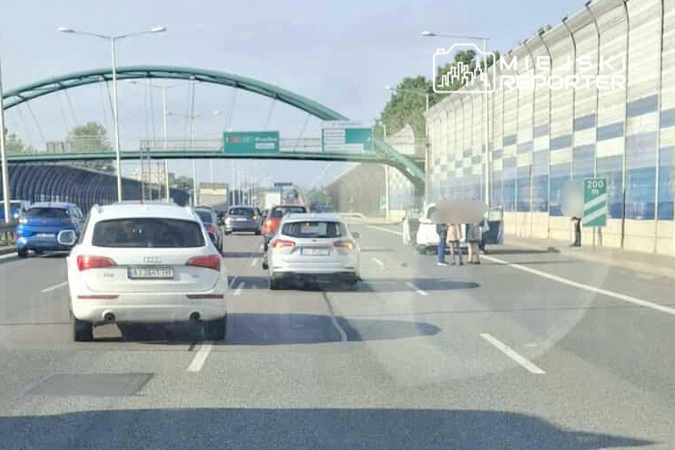 Na jezdni zatrzymane są dwa samochody, a obok nich kilka osób rozmawia przy krawężniku. W tle widoczny most i znaki drogowe.