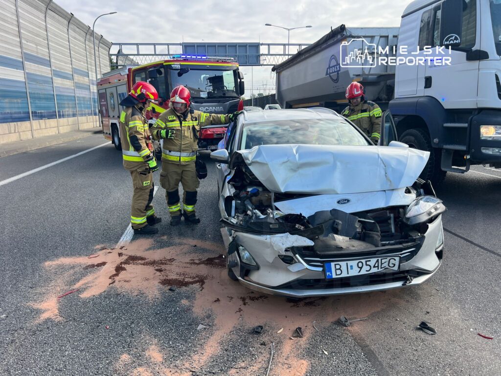 Dwaj strażacy w mundurach omawiają uszkodzone auto na drodze, obok widać woz strażacki. Na jezdni plama płynów.