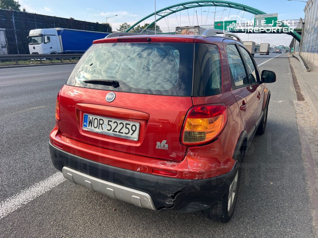 Czerwony samochód osobowy z uszkodzeniami tylnym porusza się wzdłuż drogi, obok ciężarówek.