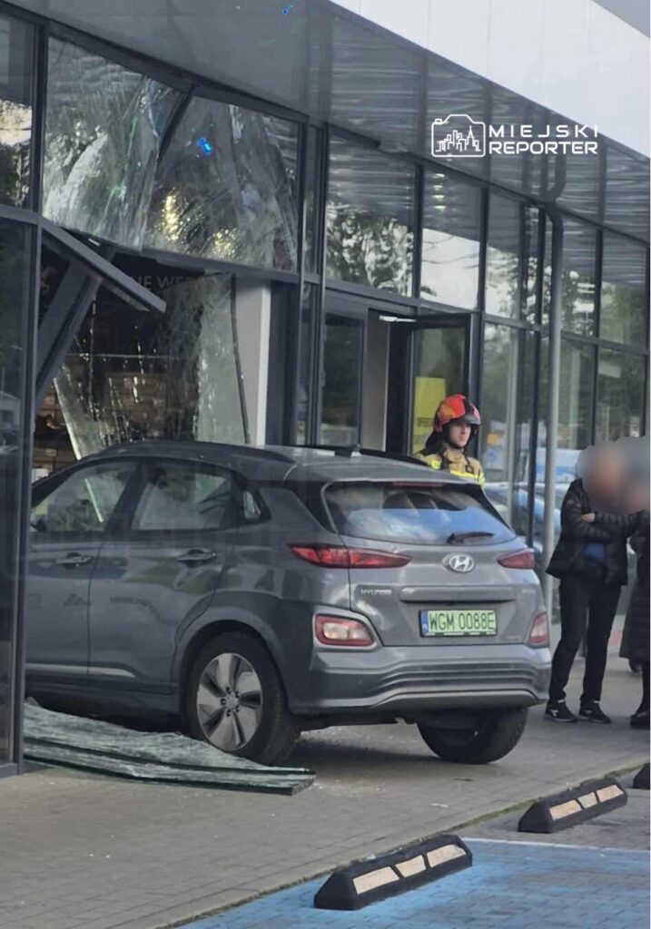 Samochód osobowy marki Hyundai wjeżdża w szklaną fasadę sklepu, obok stoi strażak w hełmie i dwie osoby.