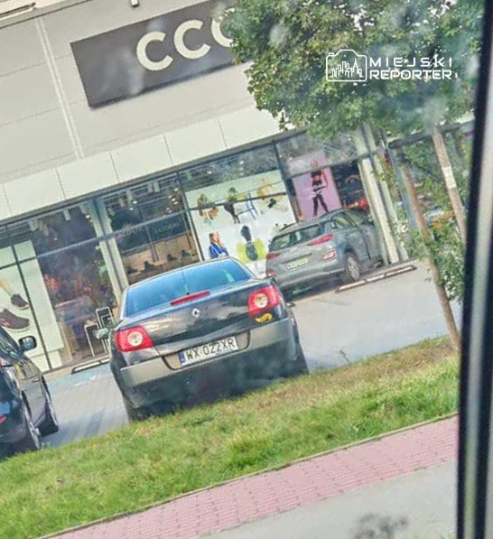 Czarny samochód zaparkowany przed nowoczesnym sklepem z dużymi witrynami, w tle widać osoby przebywające w środku.