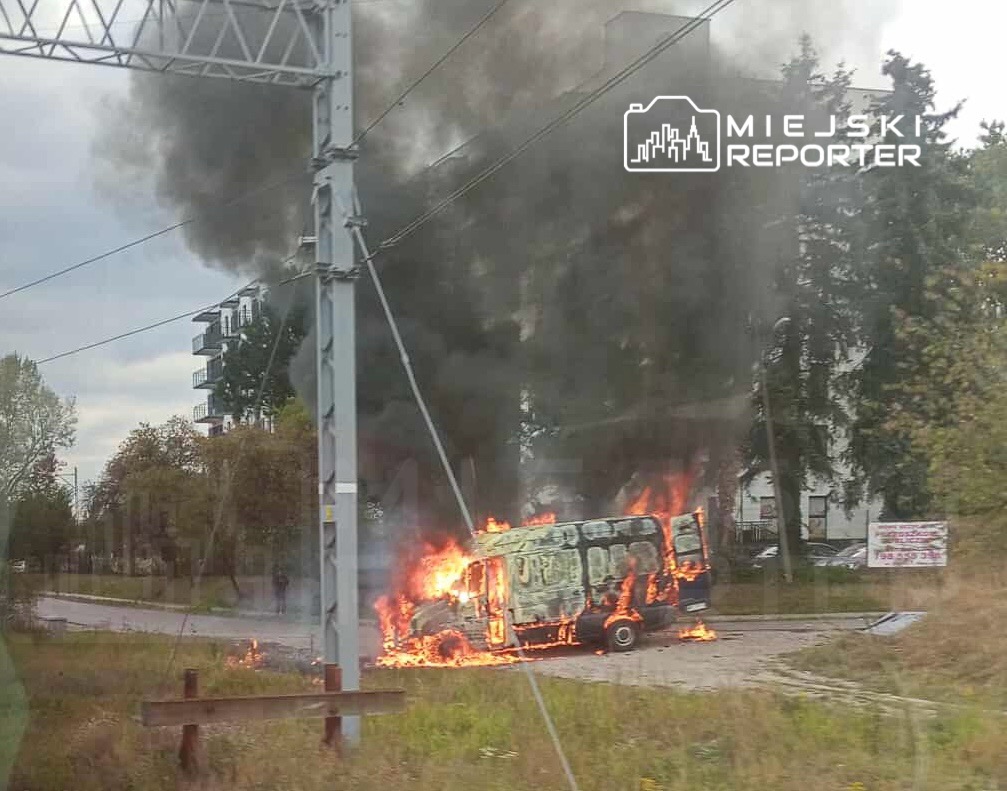 Zaparkowany pojazd płonący na poboczu drogi, w tle widoczne budynki mieszkalne i gęsta chmura dymu.