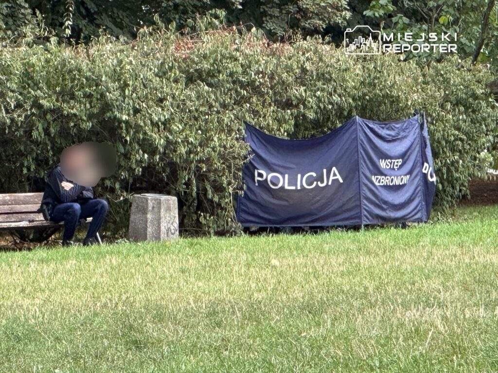 Mężczyzna siedzący na ławce przy oznakowanym namiocie policyjnym w parku, z zielonymi krzewami w tle.