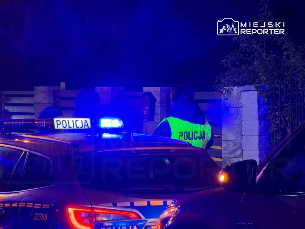 Policjanci w odblaskowych kamizelkach rozmawiają przy radiowozie w nieoświetlonym otoczeniu.