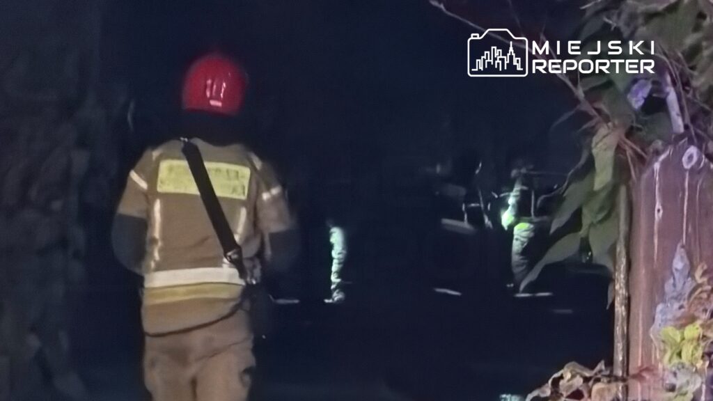 Strażak w hełmie i mundurze przeszukuje ciemne pomieszczenie, oświetlając teren latarką.