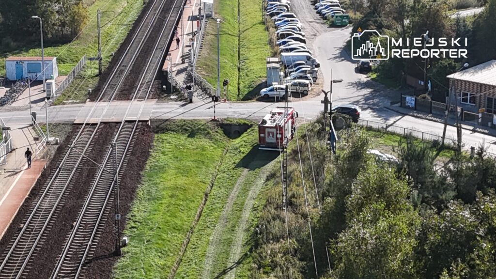 W tle torów kolejowych stoi wóz strażacki obok przejazdu, a w oddali widać zaparkowane samochody.