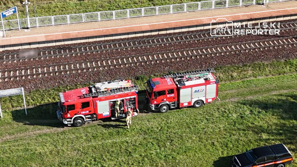 Dwa wozy strażackie zaparkowane na trawie obok torów kolejowych, strażacy sprawdzają sprzęt.