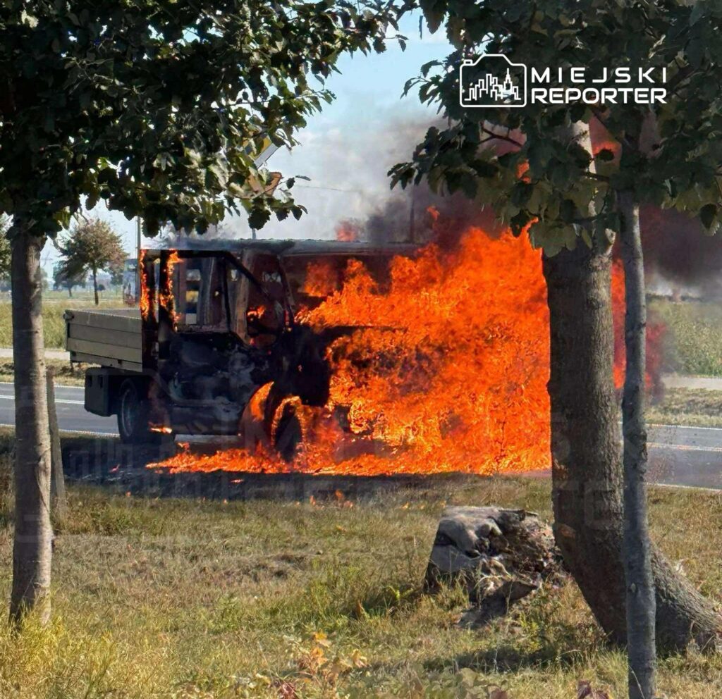 Pojazd dostawczy w płomieniach przy drodze, otoczony dymem i zielenią drzew.