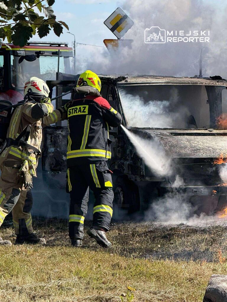 Strażacy gaszą płonący pojazd na poboczu drogi, obok widoczny znak drogowy. W tle widać dym i ogień.