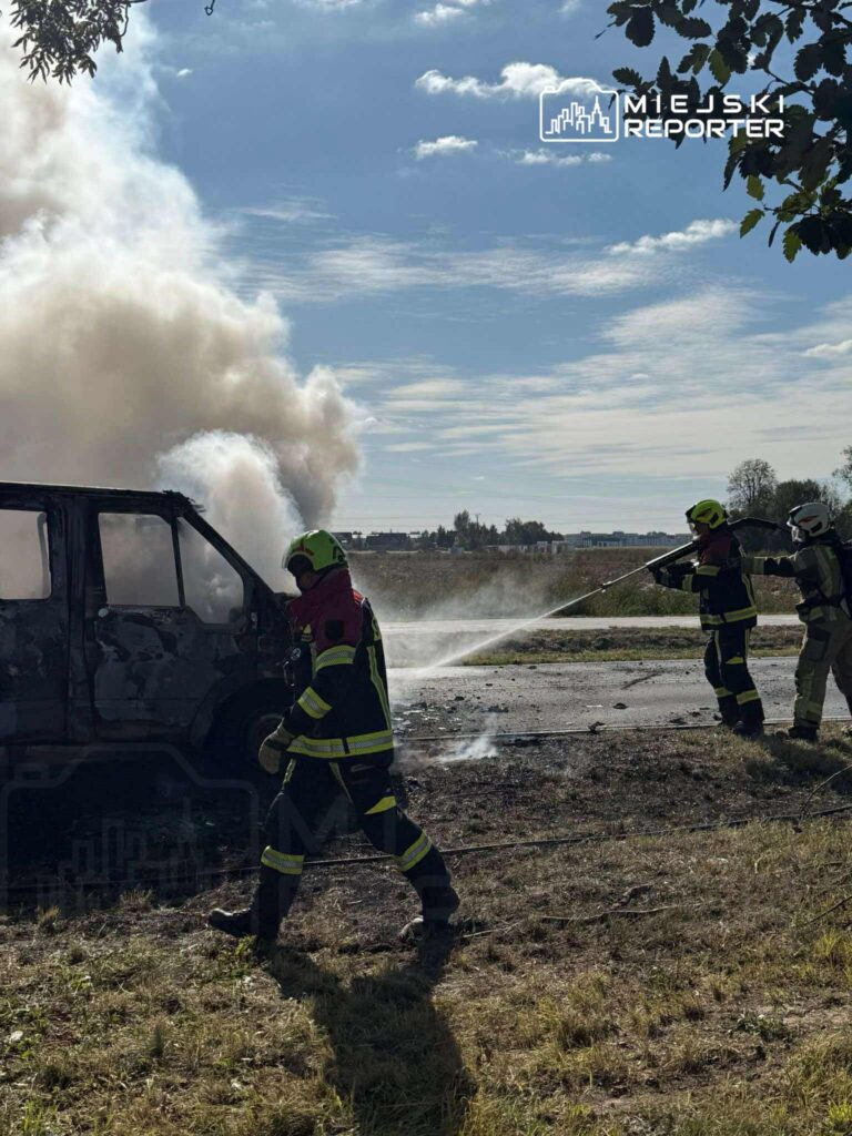 Strażacy gaszą pożar wraku samochodu na polnej drodze, w tle widoczne kłęby dymu i błękitne niebo.
