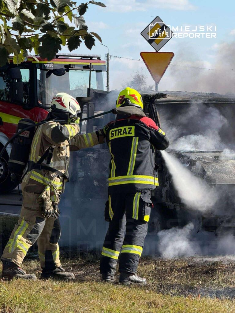 Dwaj strażacy gaszą pożar wraku samochodu, w tle widoczna strażacka ciężarówka i znak drogowy.