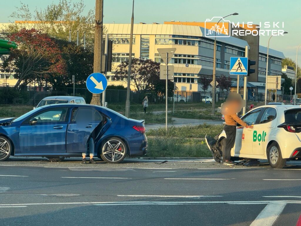 Kierowca w brązowej bluzie rozmawia z pasażerem w niebieskim samochodzie po kolizji na drodze.