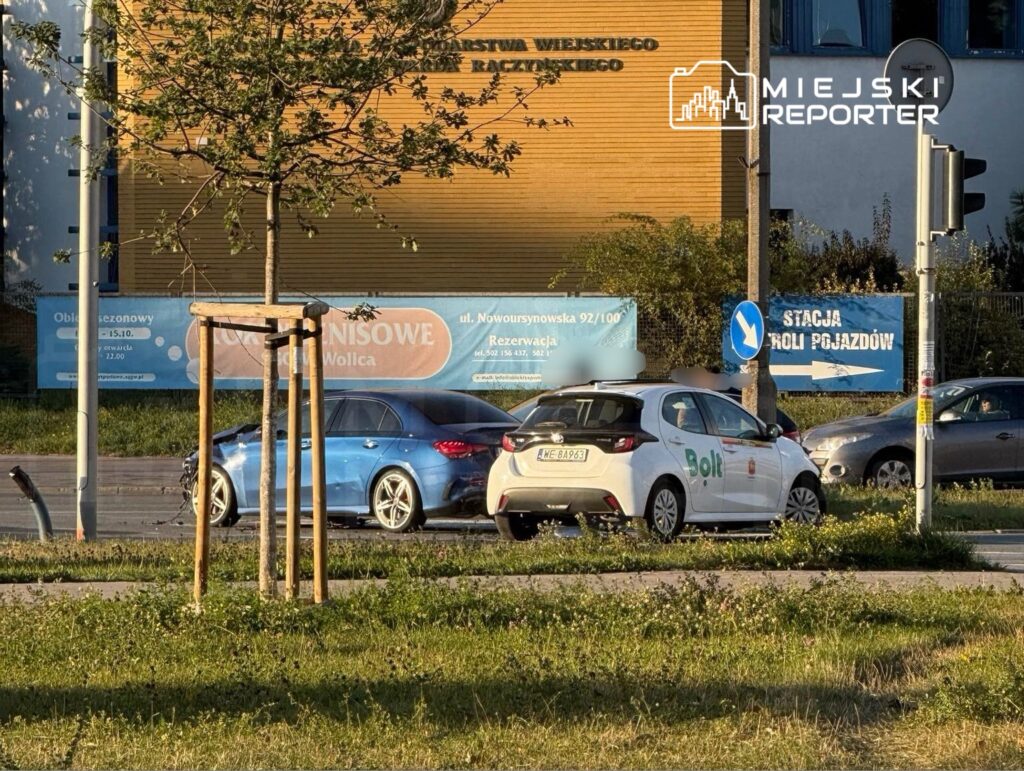 Biały samochód osobowy z logo "Bolt" zatrzymany na rondzie obok niebieskiego pojazdu, w tle billboardy i znaki drogowe.