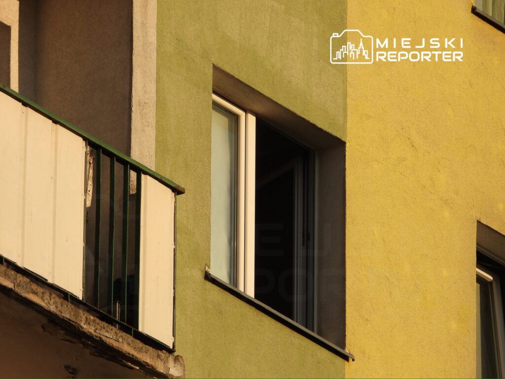 Balkon z białą balustradą i zieloną ścianą, z otwartym oknem na żółtej elewacji budynku.