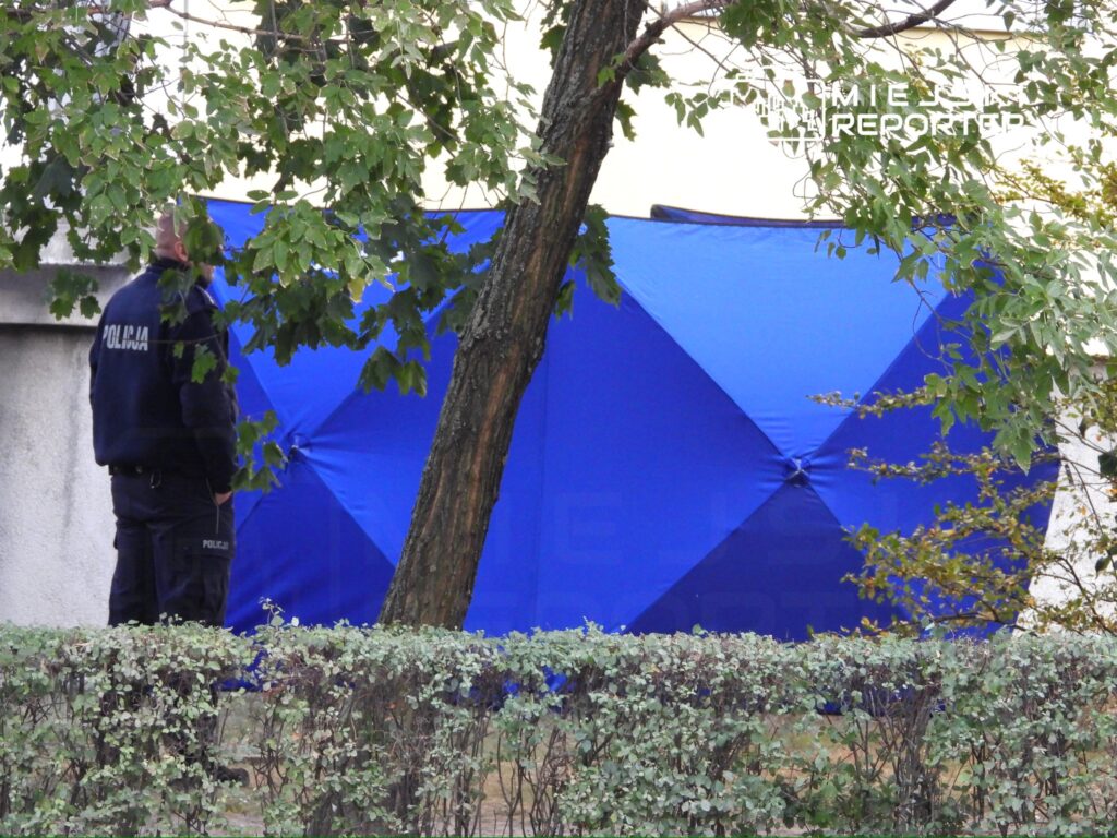 Policjant w mundurze stoi obok niebieskiego namiotu na terenie otoczonym krzewami.