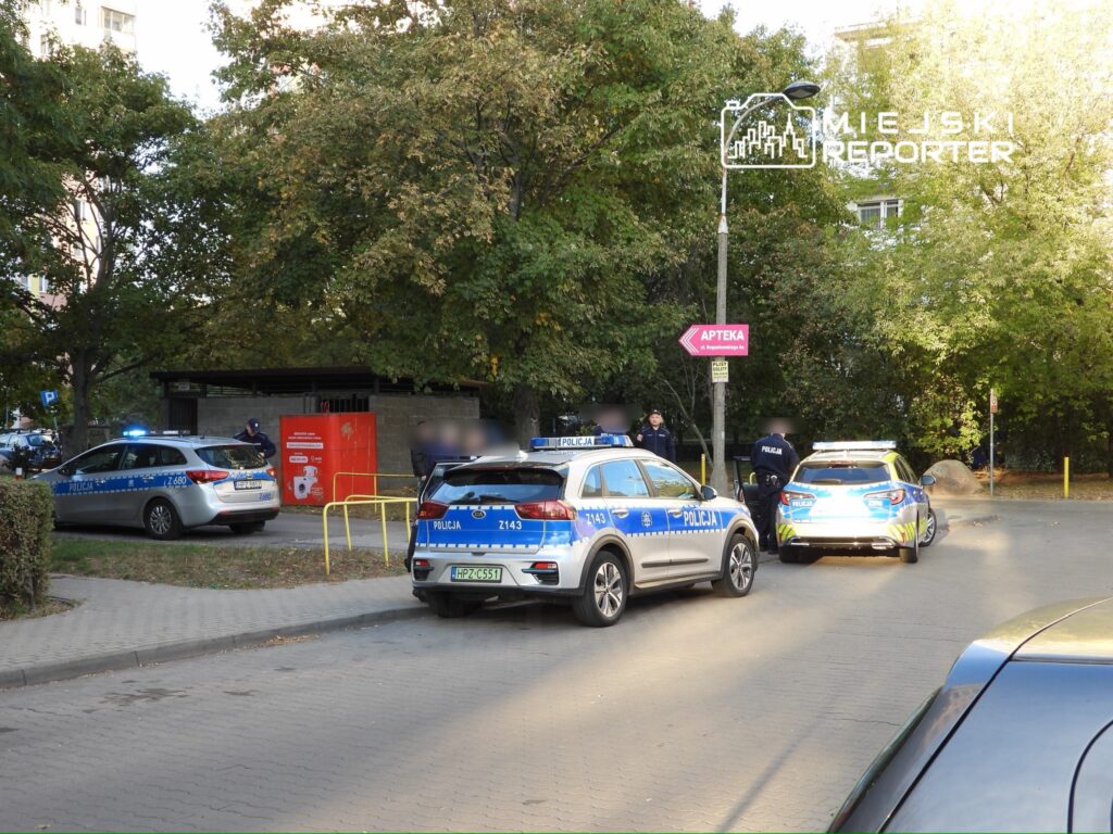Grupa policjantów rozmawia obok dwóch radiowozów na osiedlowym parkingu, w tle widoczna jest apteka.