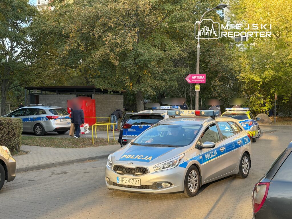 Policyjny radiowóz zaparkowany na ulicy, a funkcjonariusze prowadzą czynności w pobliżu czerwonego budynku.