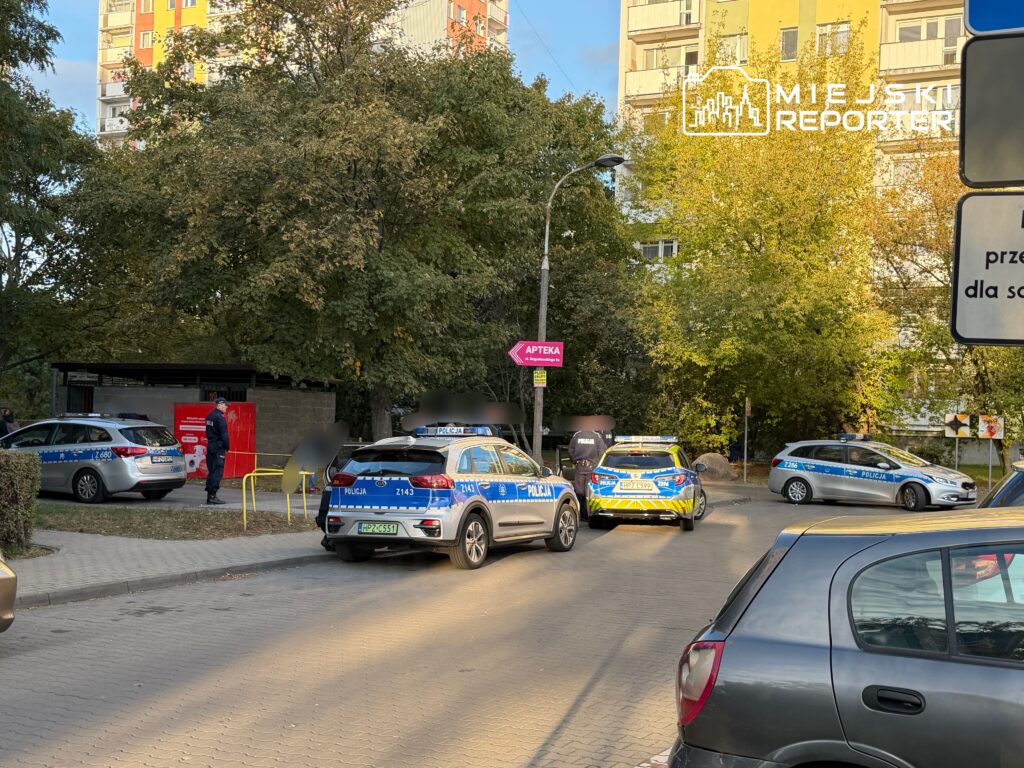 Policjanci w mundurach rozmawiają obok radiowozów na osiedlowym parkingu, w tle bloki mieszkalne.
