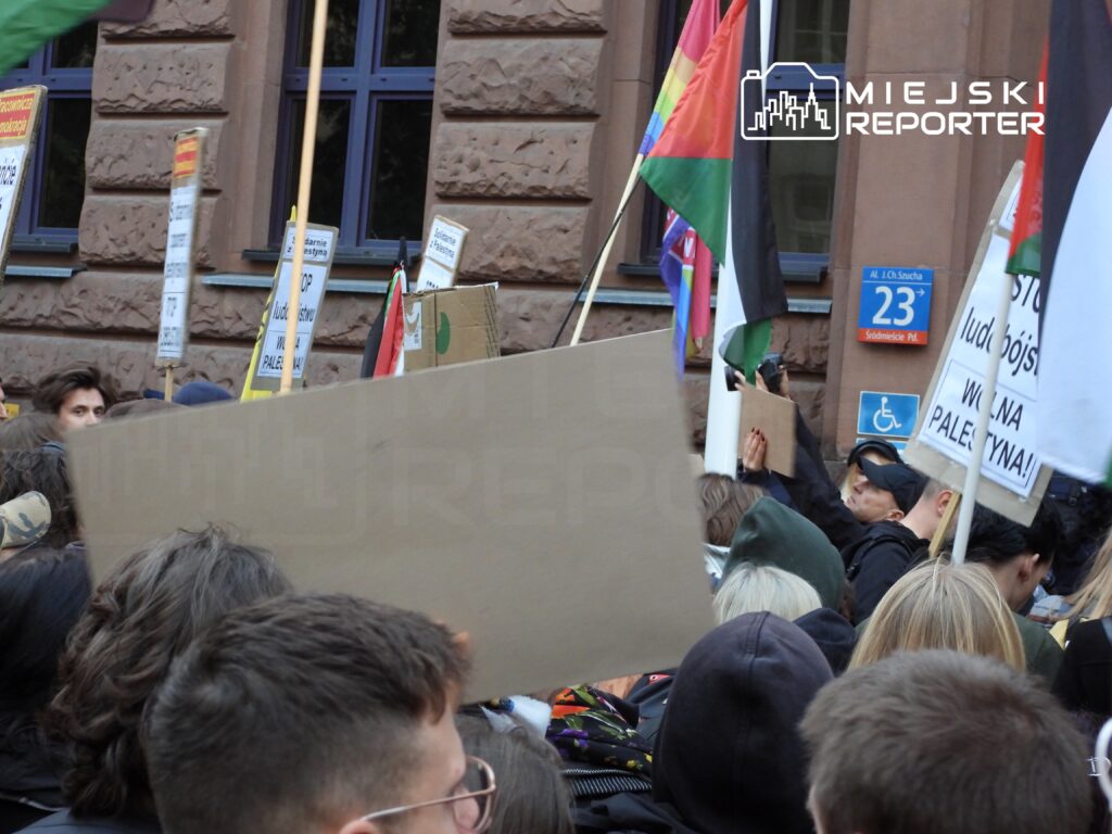 Tłum ludzi z transparentami i flagami, protestujących przed budynkiem z brązową elewacją.