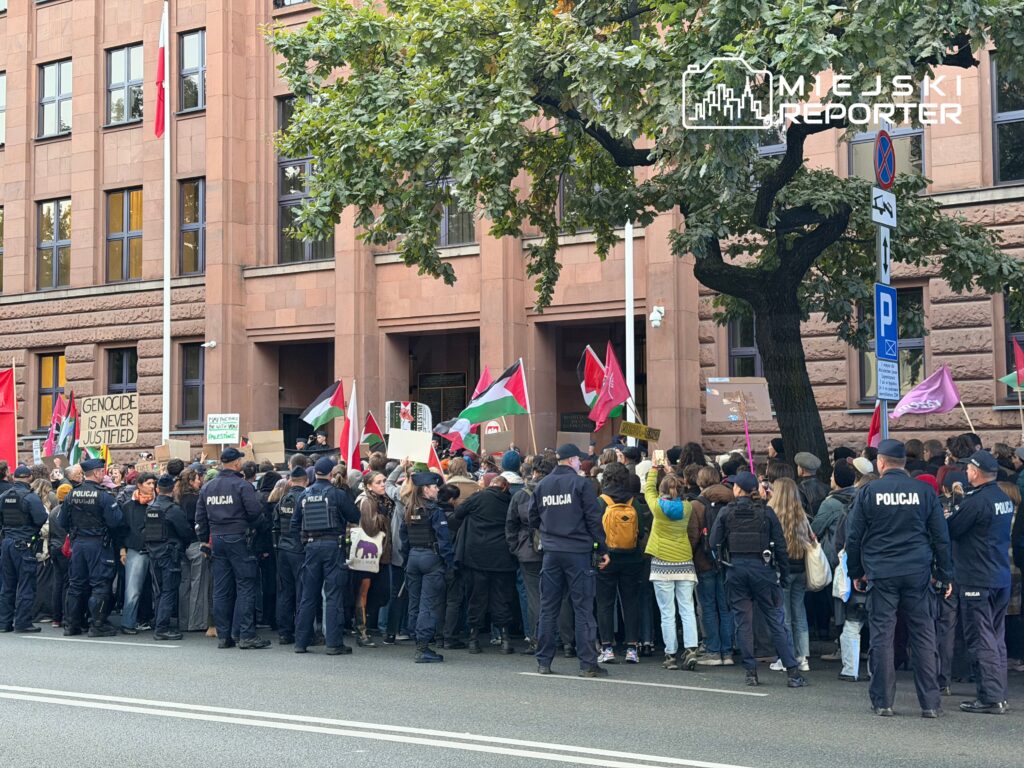 Tłum demonstrantów z transparentami i flagami zgromadzony przed budynkiem, w otoczeniu policji.