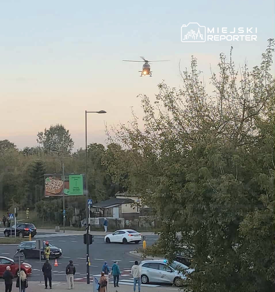 Helikopter przelatujący nad skrzyżowaniem, w tle grupka ludzi i pojazdy czekające na sygnalizację.