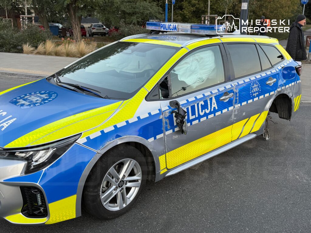 Uszkodzony radiowóz policyjny z otwartą drzwiami, zaparkowany na ulicy, z widocznymi śladami uszkodzeń na nadwoziu.