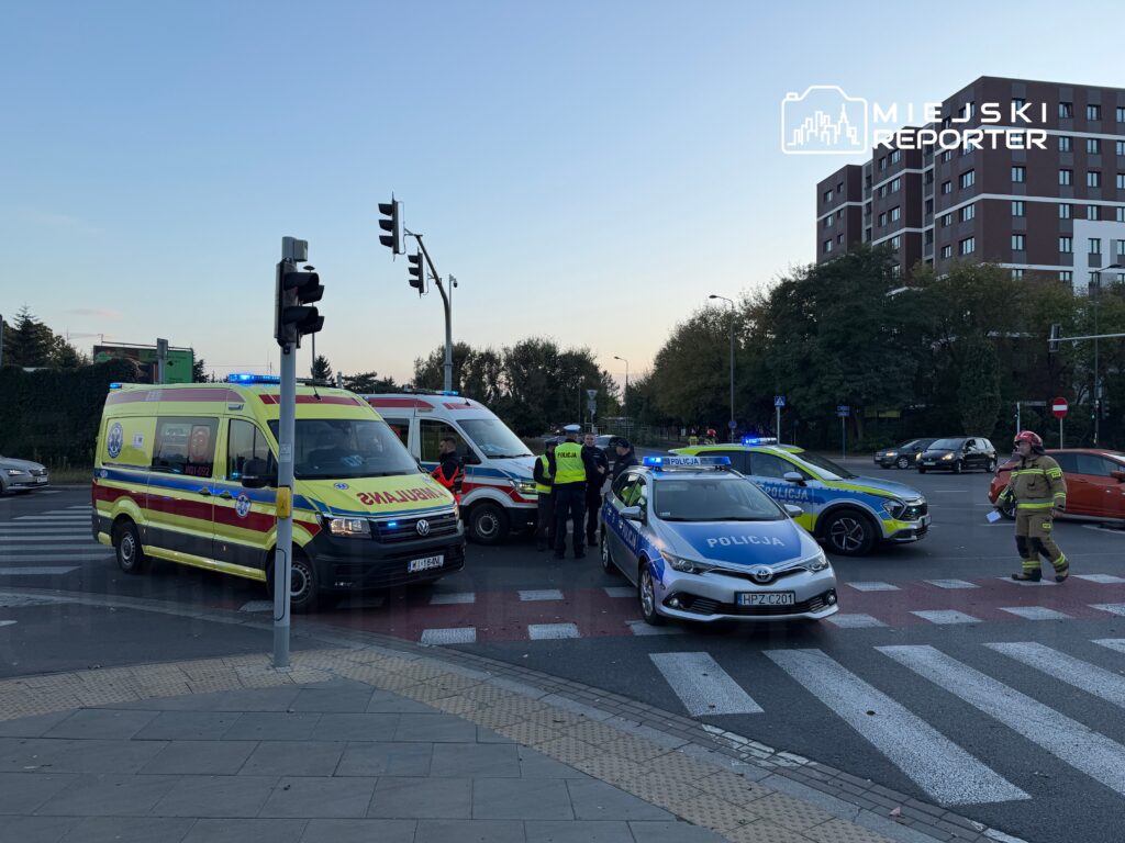 Karetka pogotowia oraz radiowozy policji blokują skrzyżowanie, podczas gdy strażak i funkcjonariusze rozmawiają z kierowcami.