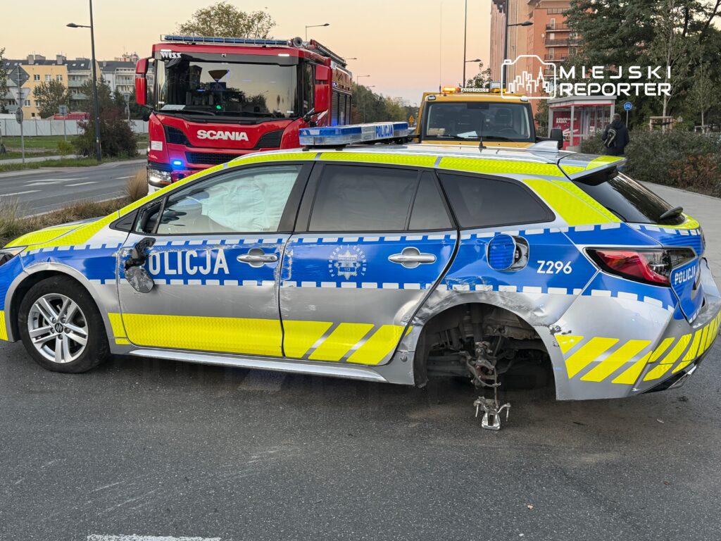 Uszkodzony radiowóz policyjny z brakiem jednego koła, stojący na jezdni obok wozu strażackiego.