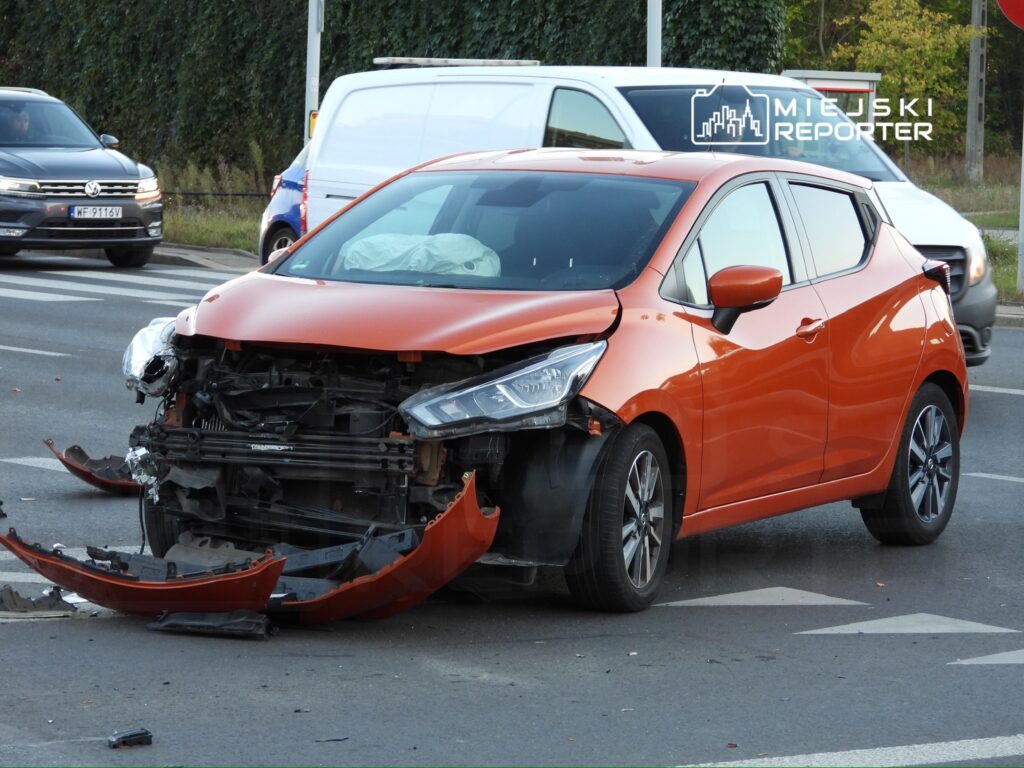 Zniszczony pomarańczowy samochód osobowy stoi na skrzyżowaniu, z uszkodzoną przednią częścią i brakującym zderzakiem.
