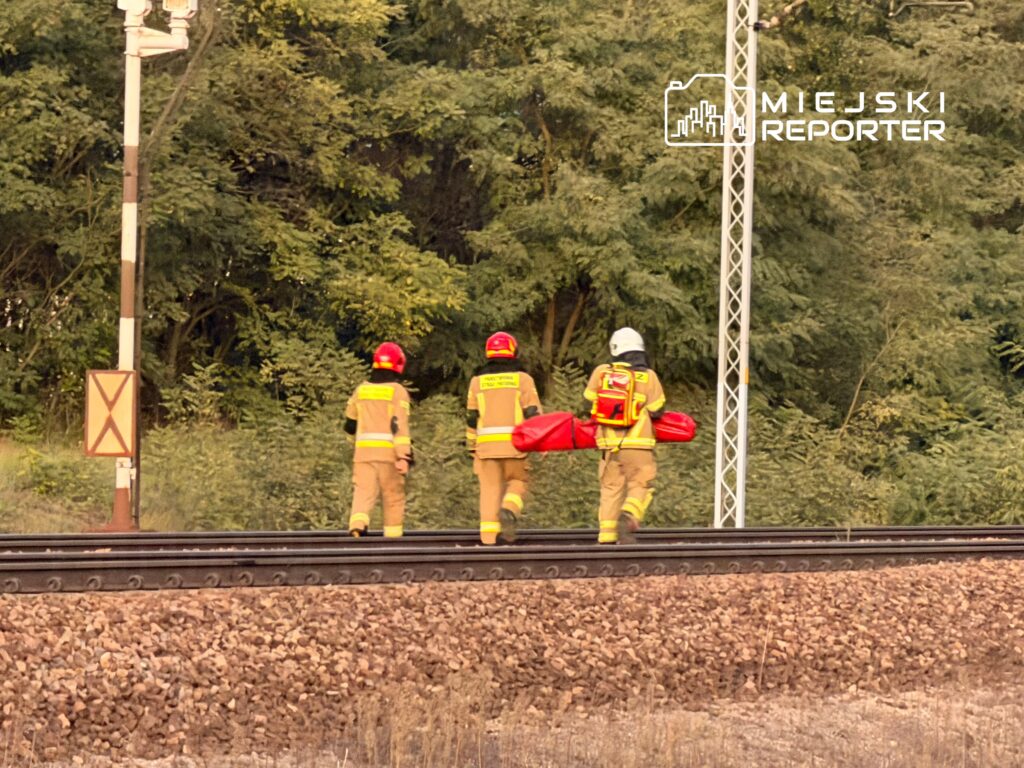 Trzech strażaków w żółtych mundurach idzie wzdłuż torów kolejowych, niosąc czerwoną nosze.
