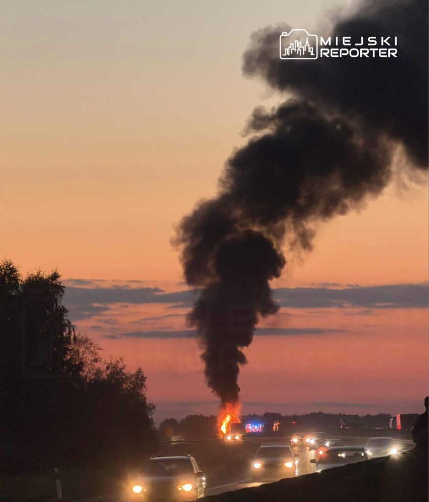 Czarna chmura dymu unosi się nad płonącym pojazdem przy drodze, w tle zachód słońca i przejeżdżające samochody.