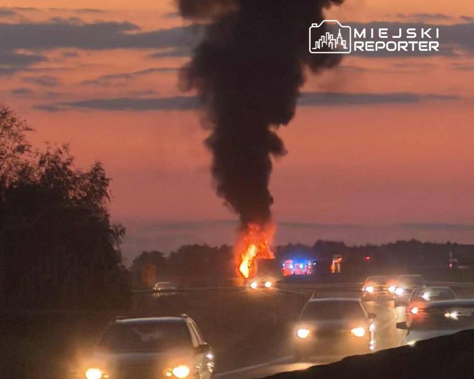 Pojazdy przejeżdżają obok płonącego wraku na drodze, w tle widoczny dym i policyjne światła.