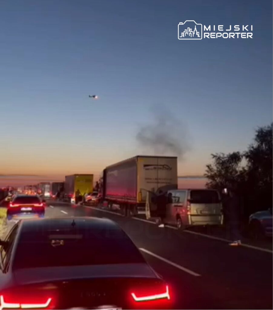 Samochody stoją w korku na drodze, a w tle unosi się dym z ciężarówki, nad którą przelatuje helikopter.
