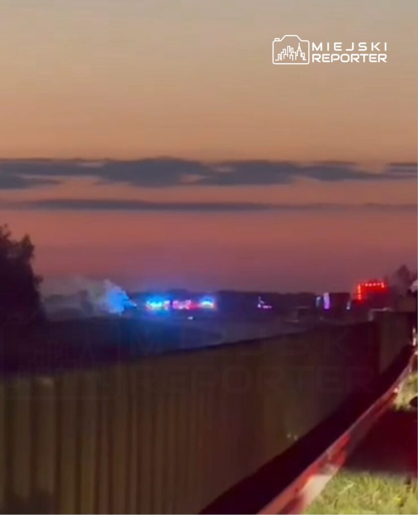 Na drodze w oddali widoczne niebieskie i czerwone światła pojazdów służb ratunkowych, w tle zachód słońca.