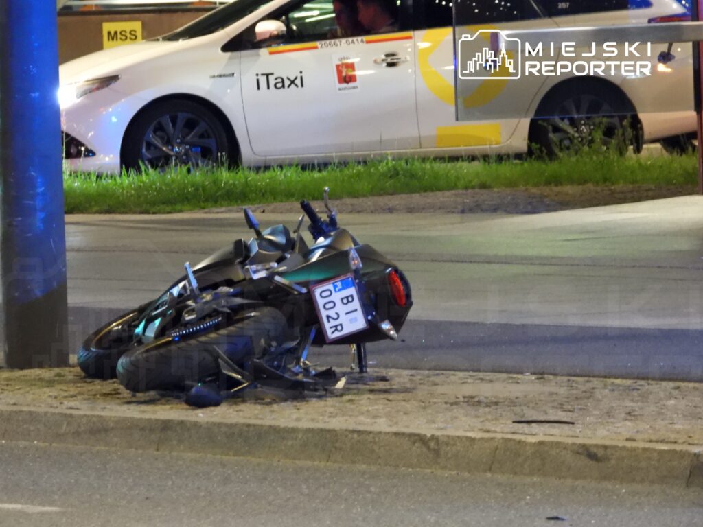 Zniszczony motocykl leży na chodniku obok słupa, a w tle widoczny jest taksówka z napisem "iTaxi".