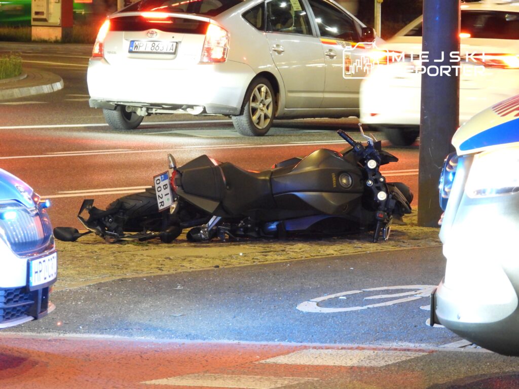 Motocykl leży przewrócony na jezdni, obok zaparkowanych samochodów, w nocy, w otoczeniu świateł policyjnych.