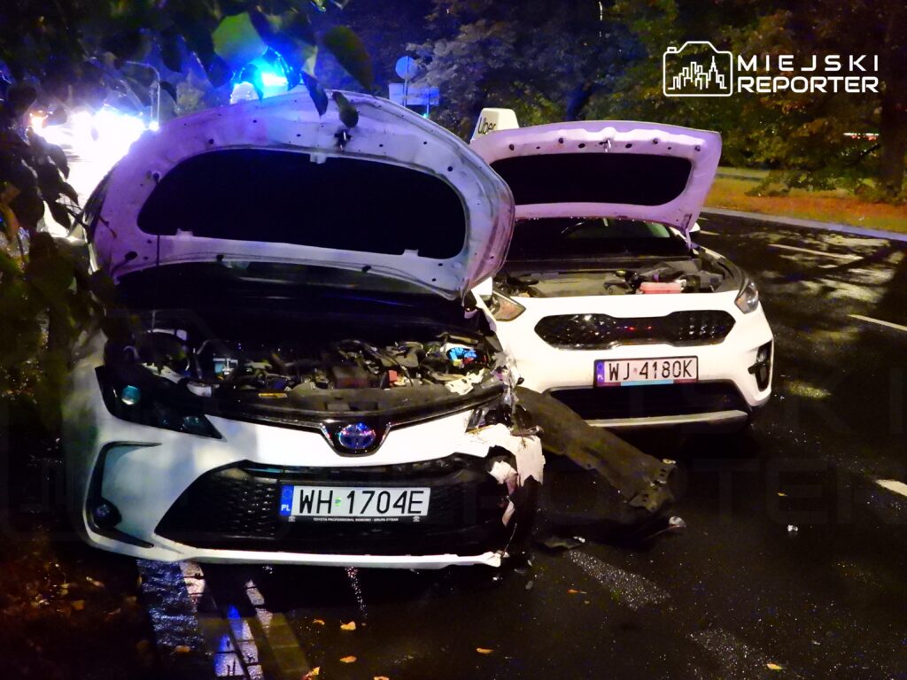 Dwa uszkodzone samochody osobowe zaparkowane na mokrej nawierzchni, z otwartymi maskami, w nocy.