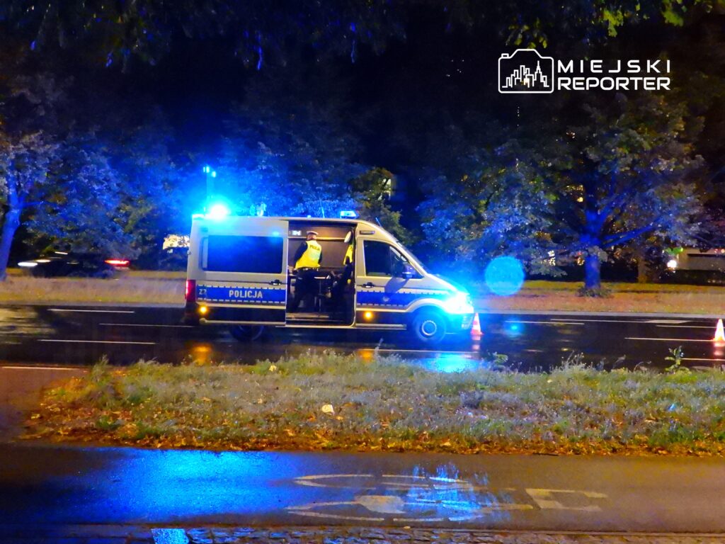 Policyjny radiowóz z włączonymi sygnałami świetlnymi stoi na poboczu drogi w nocy, obok widoczne są pachołki.