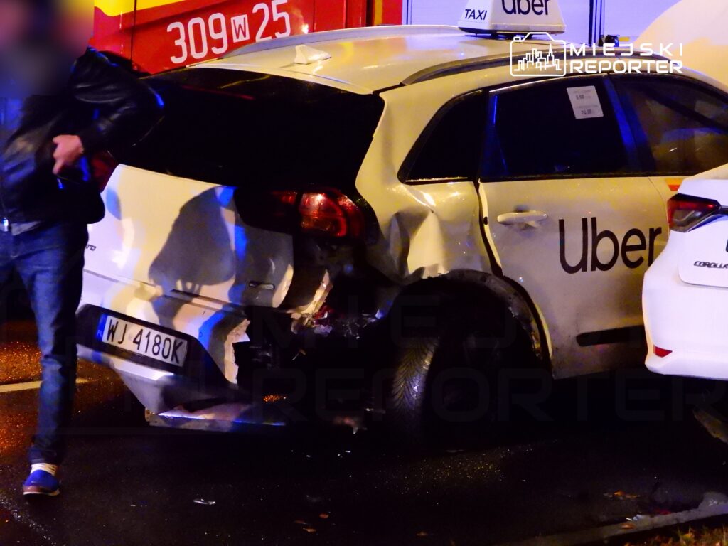 Mężczyzna w czarnej kurtce stoi obok uszkodzonego samochodu Ubera na mokrej nawierzchni ulicy.