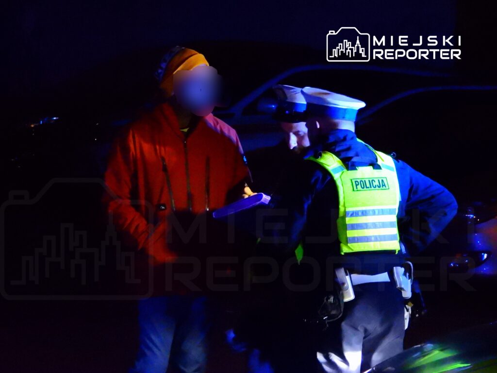 Mężczyzna w pomarańczowej kurtce rozmawia z policjantami na ulicy w nocy, przy oświetleniu radiowozu.