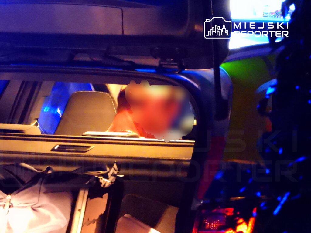 Osoba siedząca w bagażniku samochodu, otoczona niebieskim światłem policyjnym.