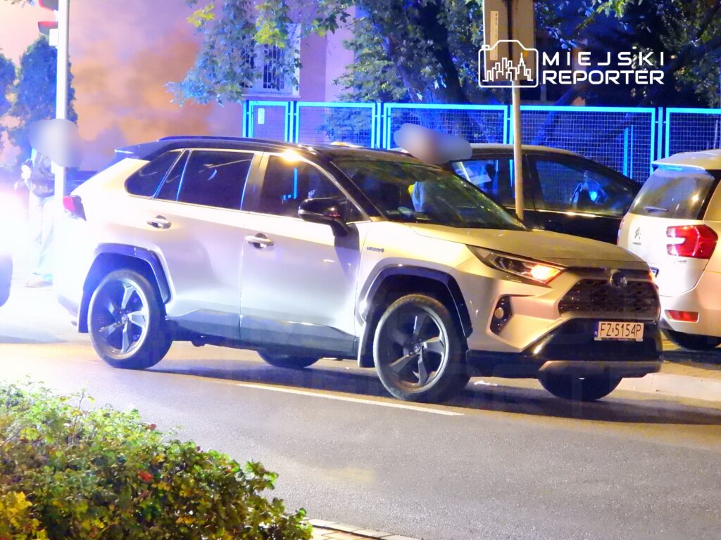 Biały SUV zaparkowany na ulicy w nocy, obok stojącej postaci oraz pojazdów w tle.