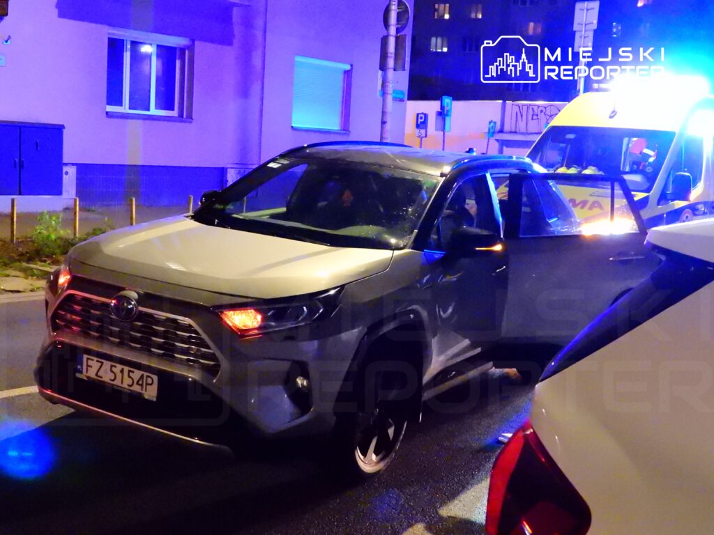 Samochód osobowy zaparkowany na ulicy nocą, obok karetki pogotowia z włączonymi sygnałami świetlnymi.