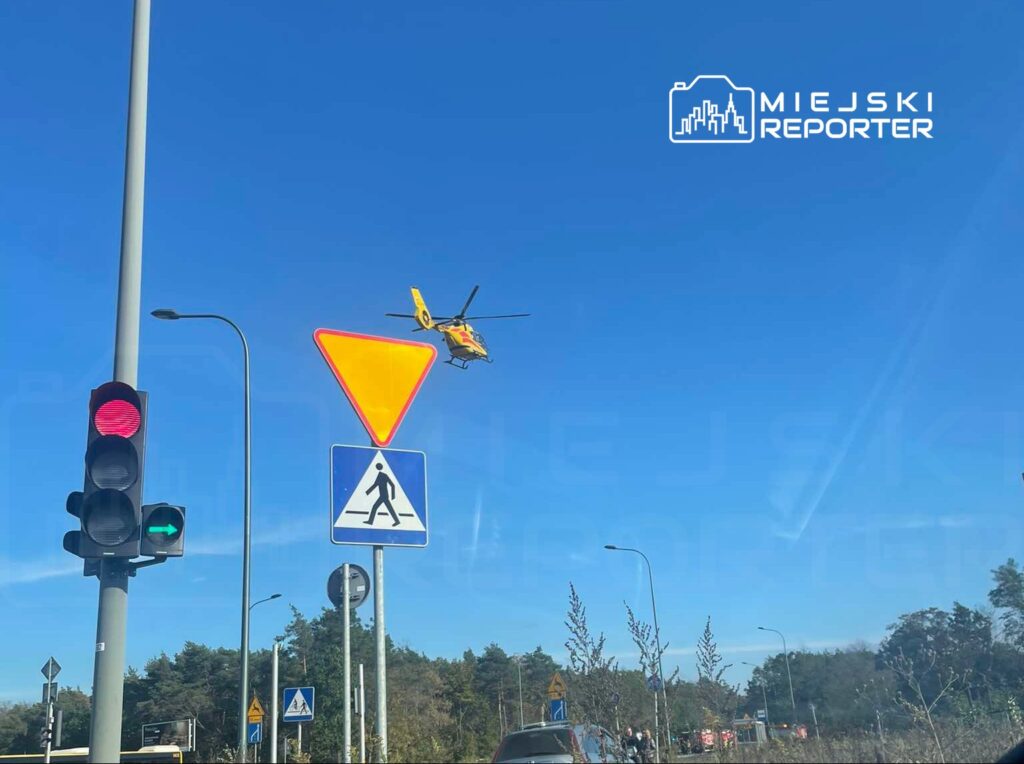 Śmigłowiec ratunkowy przelatujący nad sygnalizacją świetlną w słoneczny dzień, w tle zieleń drzew.