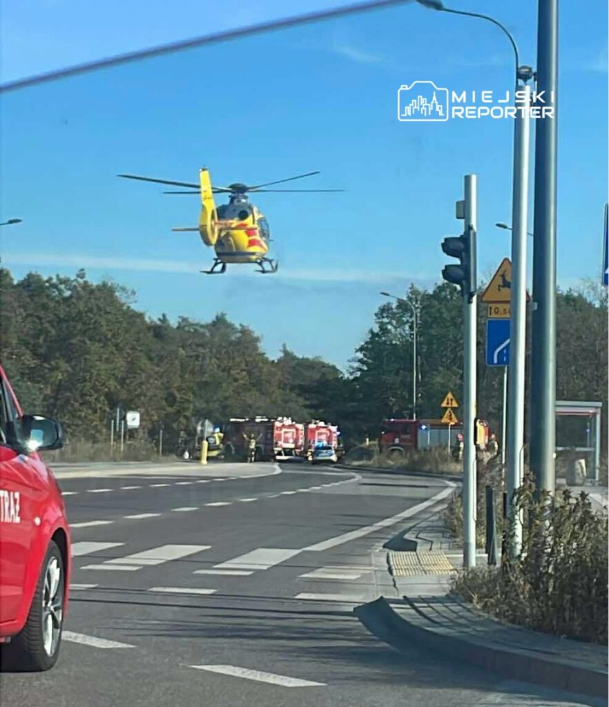 Żółty helikopter ratunkowy unosi się nad drogą, obok której stoją pojazdy straży pożarnej.