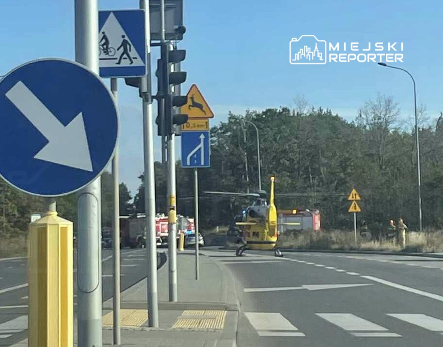 Śmigłowiec ratunkowy ląduje na poboczu drogi, obok sygnalizacji świetlnej i oznaczeń drogowych.