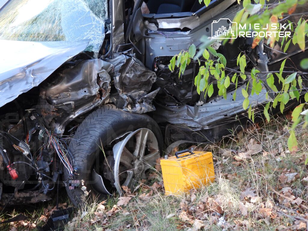 Zniszczony samochód leży w zaroślach, z odsłoniętymi przewodami elektrycznymi i uszkodzoną felgą, obok żółta skrzynka.