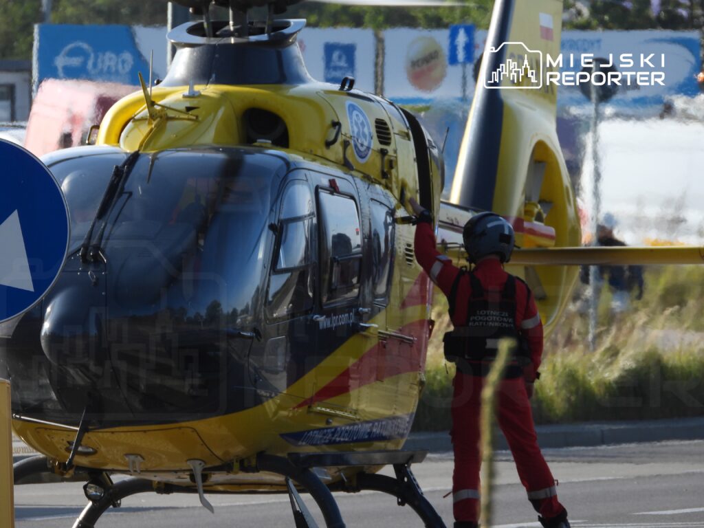 Pilot w czerwonym kombinezonie z logo Lotniczego Pogotowia Ratunkowego otwiera drzwi helikoptera na lądowisku.