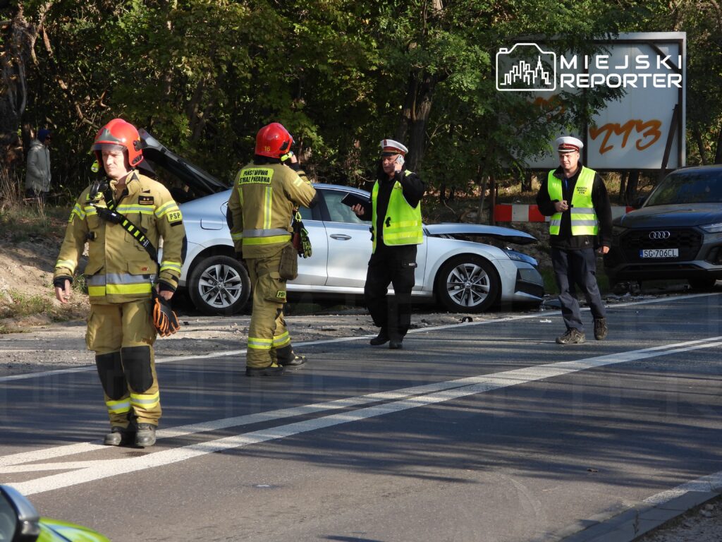 Strażacy w mundurach i hełmach pracują przy wypadku drogowym, a policjanci kierują ruchem w tle.